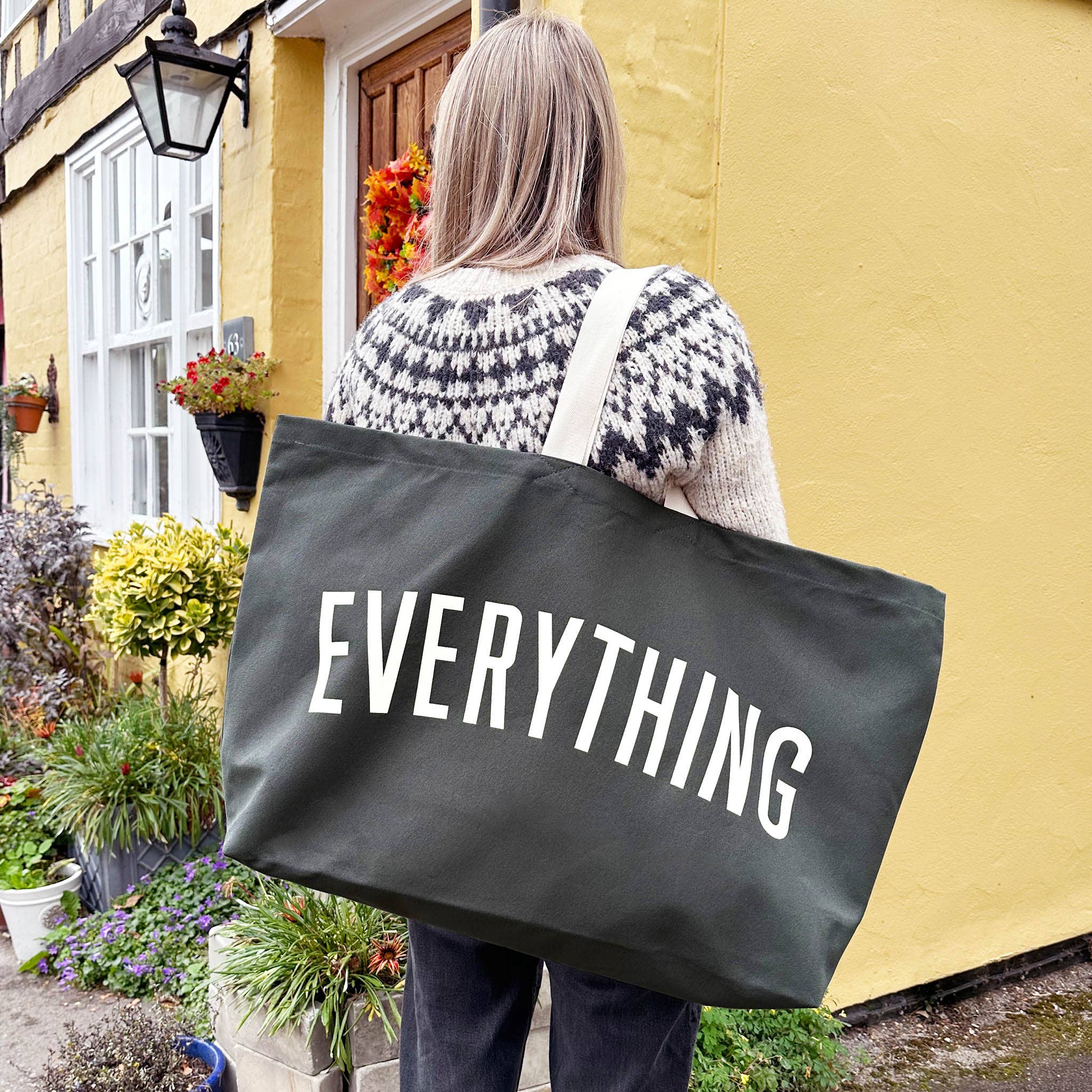 Person holding a black tote bag with 'EVERYTHING' text in front of a yellow building.