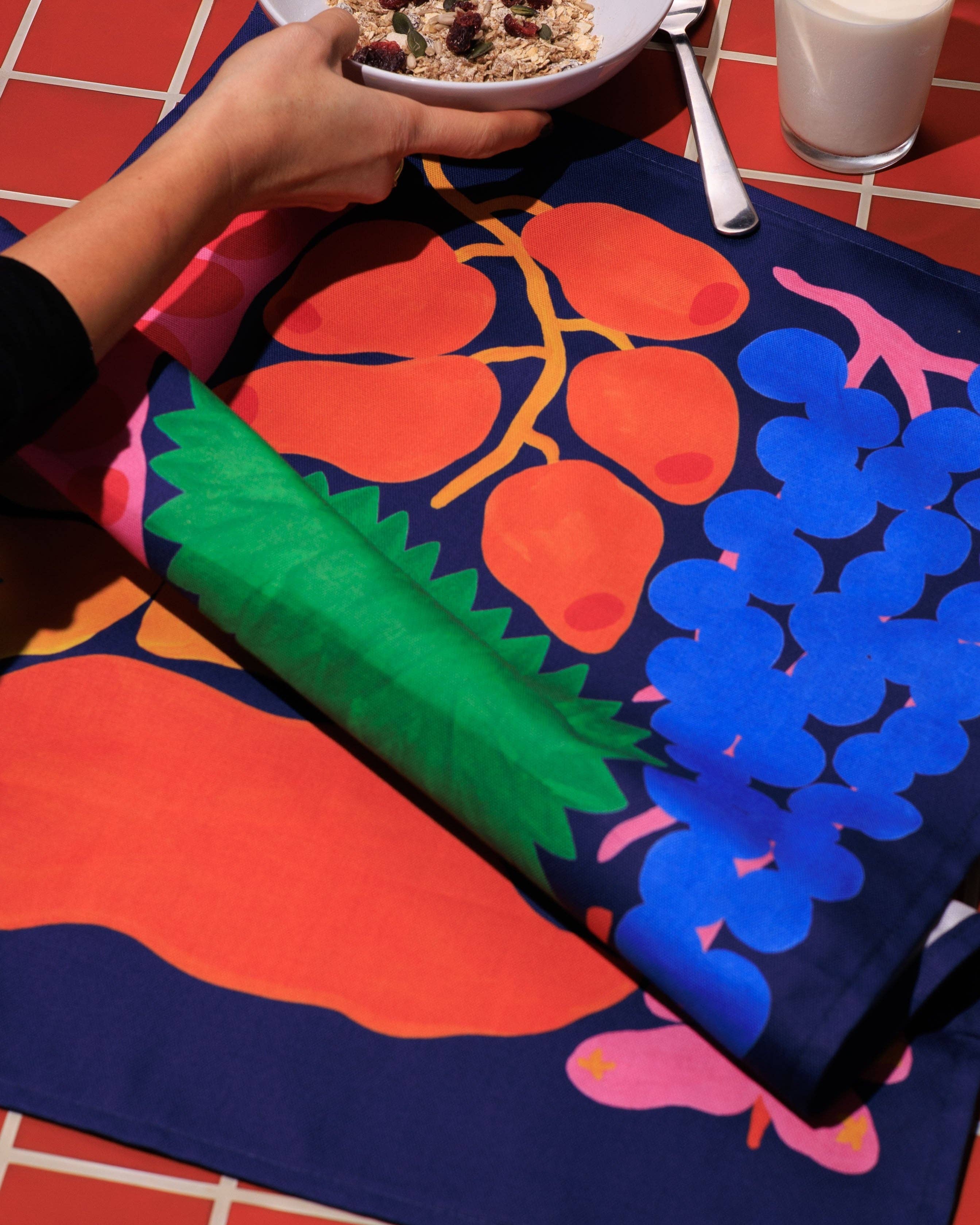Colorful tablecloth with fruit design on a table with a bowl of cereal and glass of milk.
