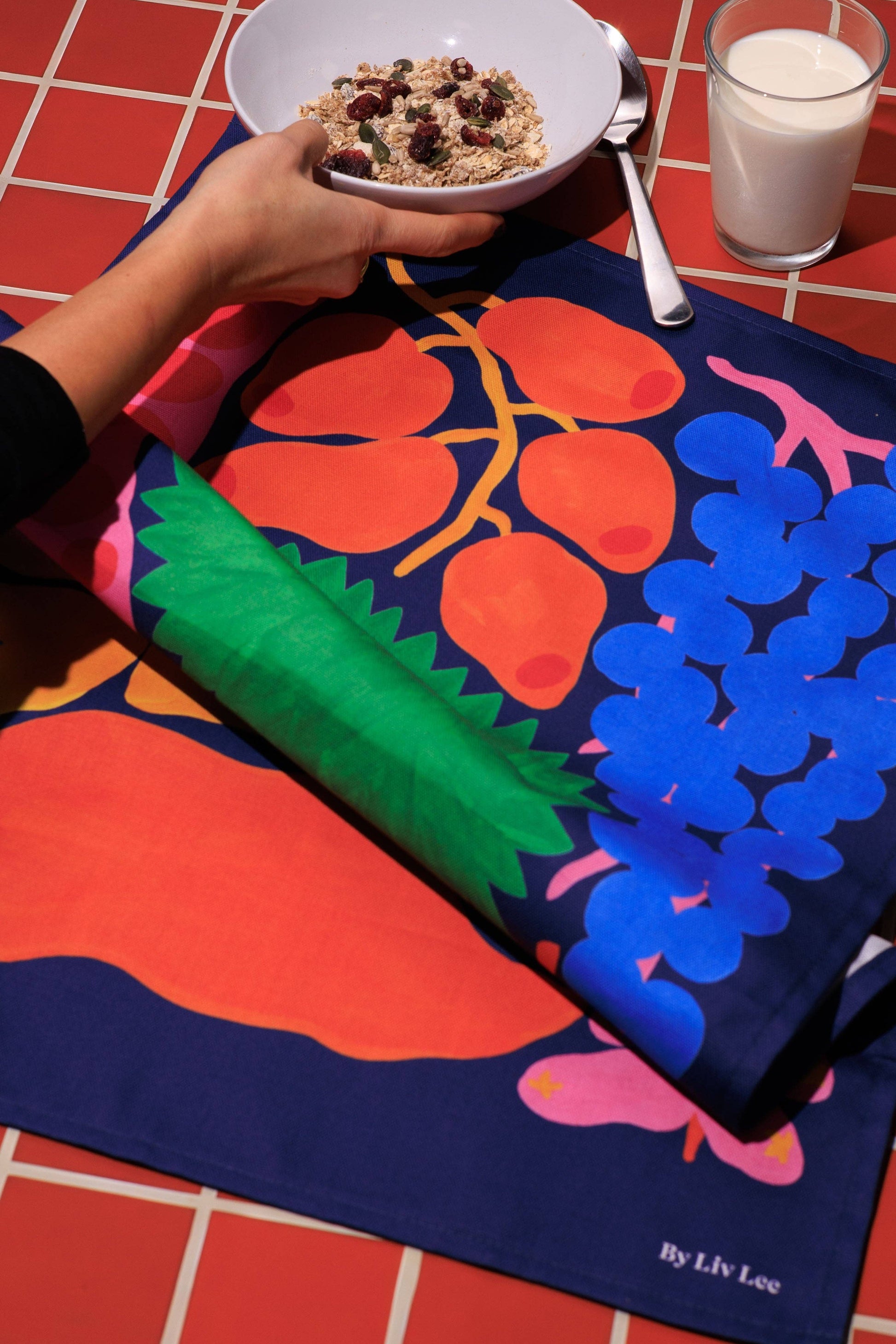 Colorful tablecloth with fruit design on a table with a bowl of cereal and glass of milk.