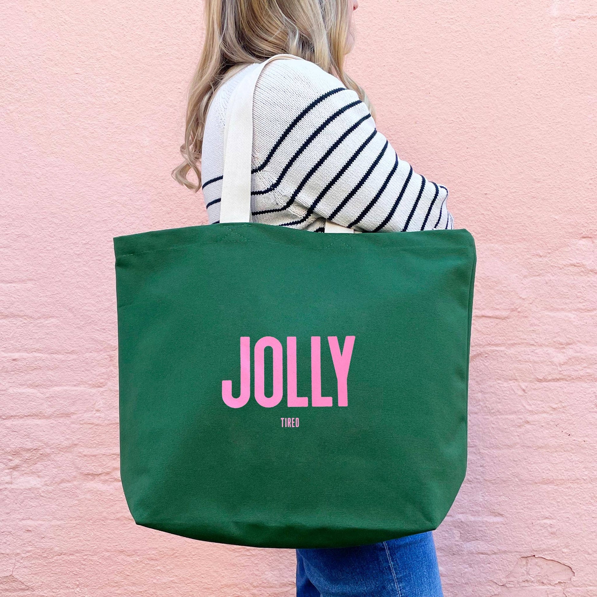 Person holding a green tote bag with 'JOLLY' printed on it against a pink background
