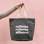 Tote bag with 'cafeine reliance alliance' text held by a hand against a pink background