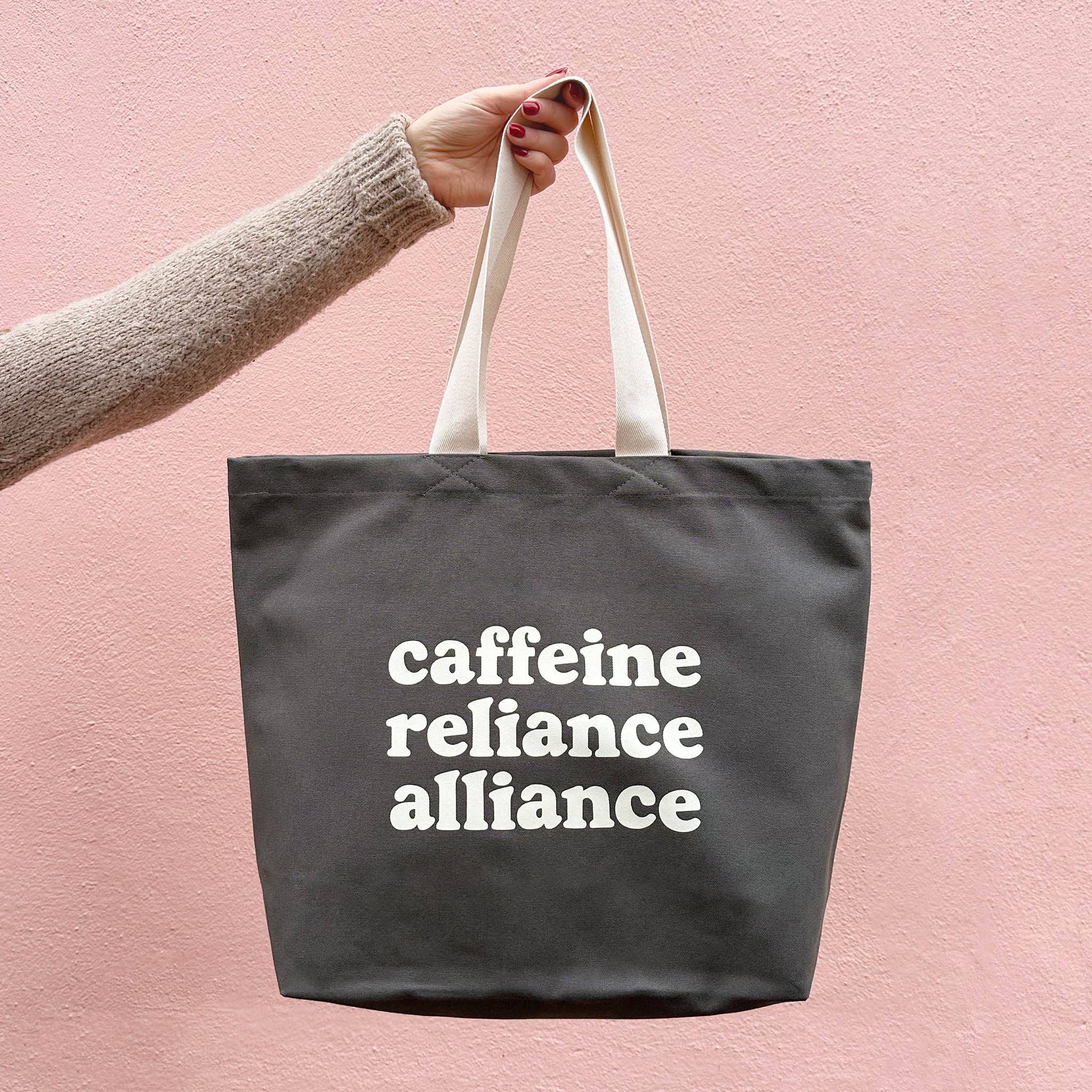 Tote bag with 'cafeine reliance alliance' text held by a hand against a pink background