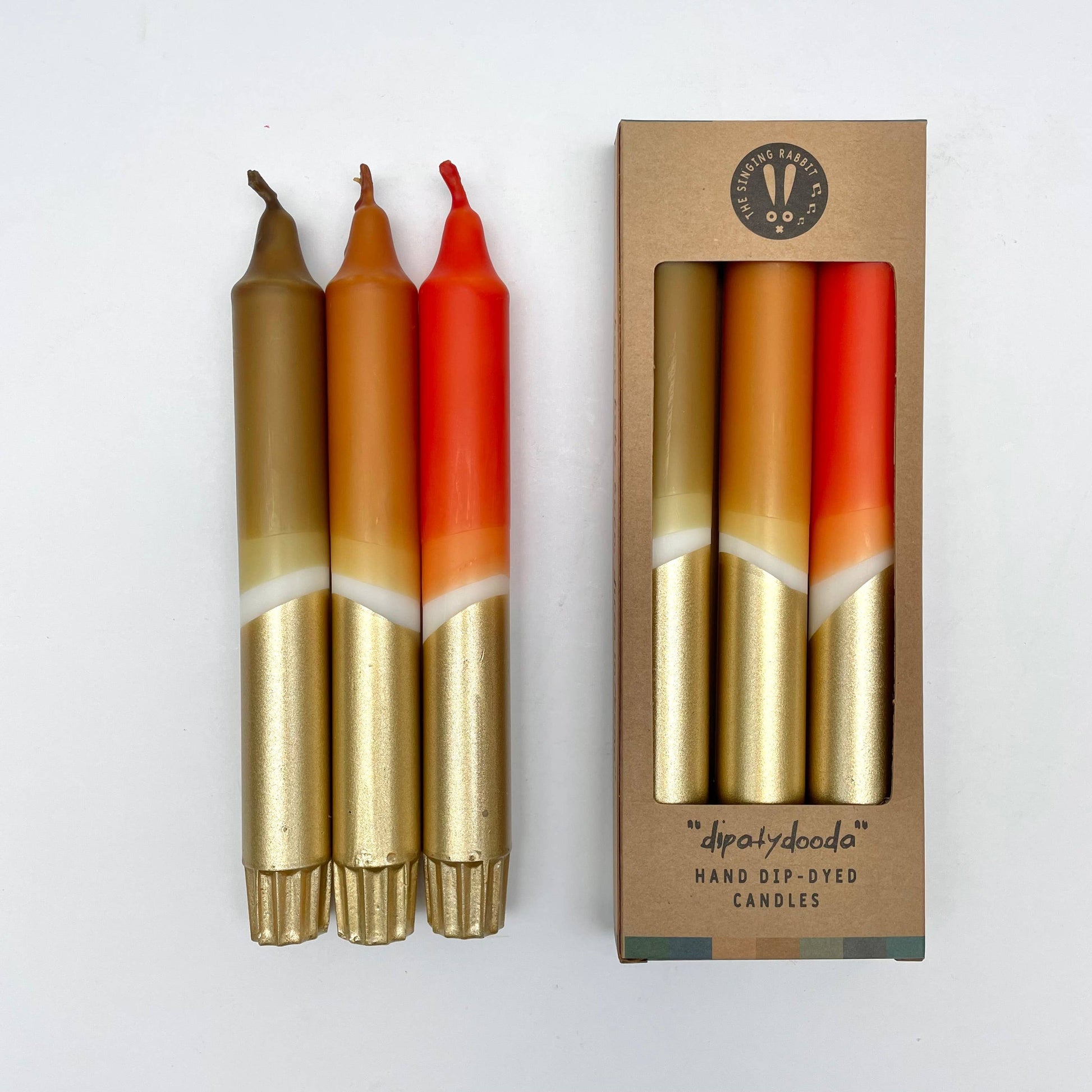 Three hand-dyed candles with gold, orange, and red colors in a cardboard box on a white background.