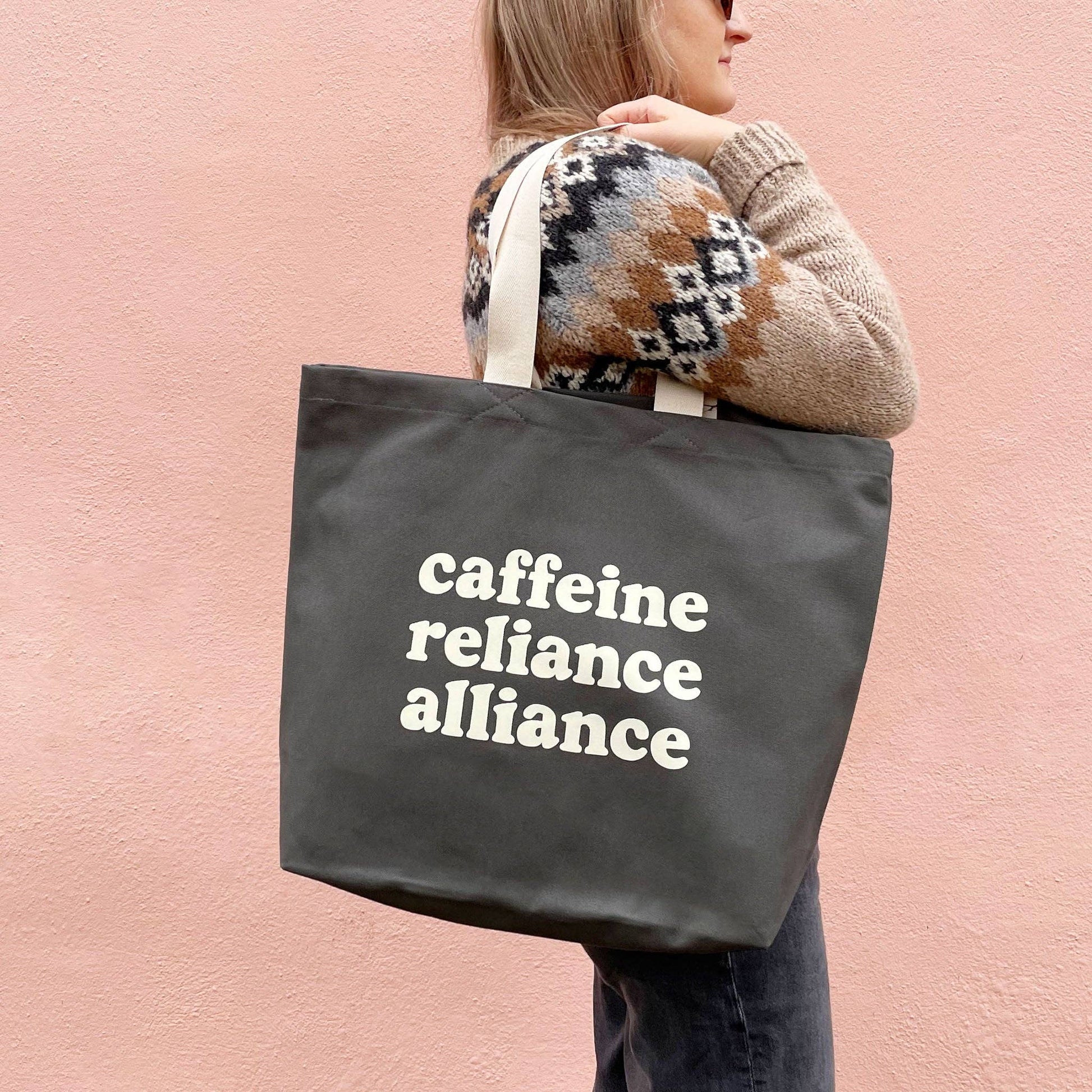 Person holding a tote bag with 'cafeine reliance alliance' text against a pink background