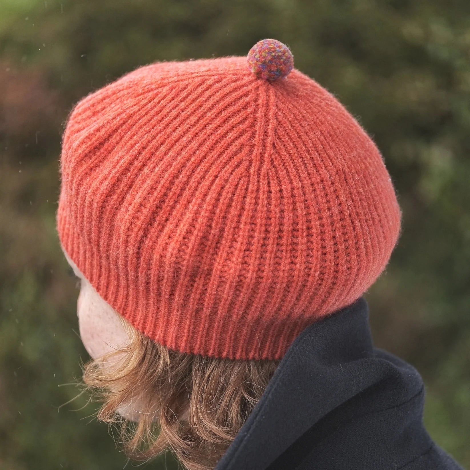 Person wearing a red knitted beanie with a pom-pom against a blurred natural background