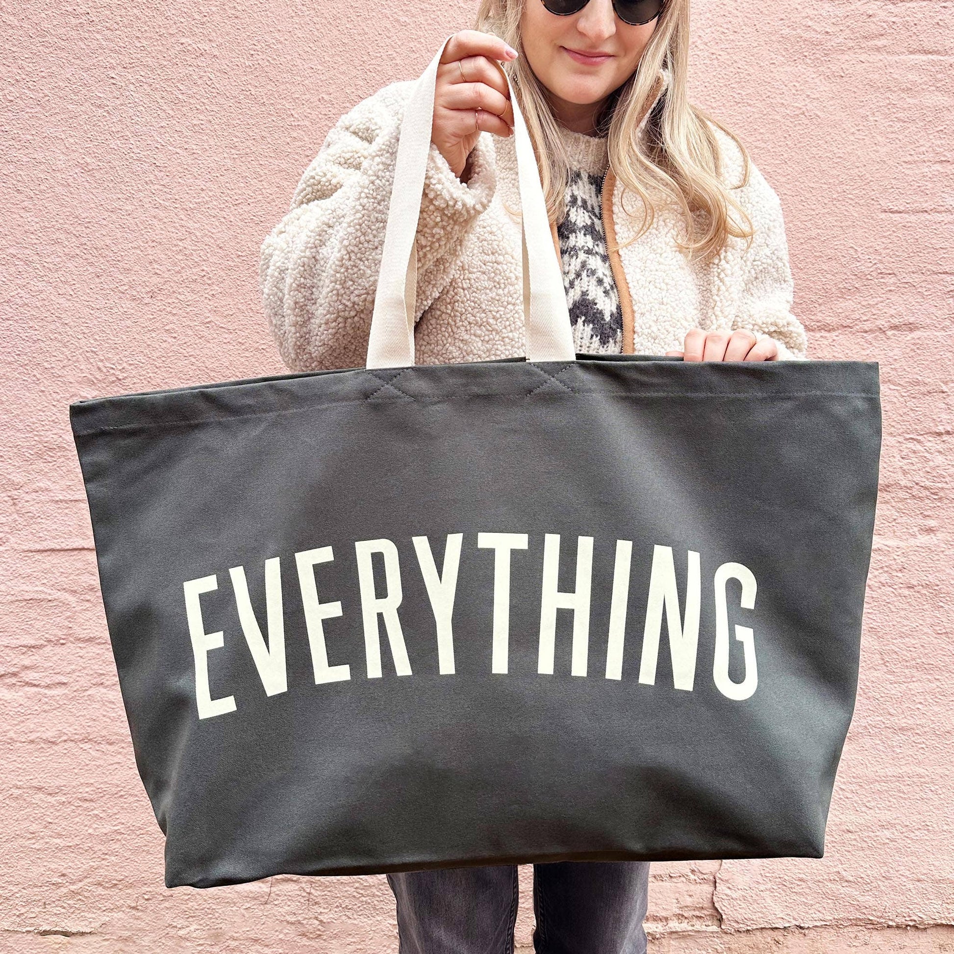 Person holding a black tote bag with 'EVERYTHING' printed on it against a pink background