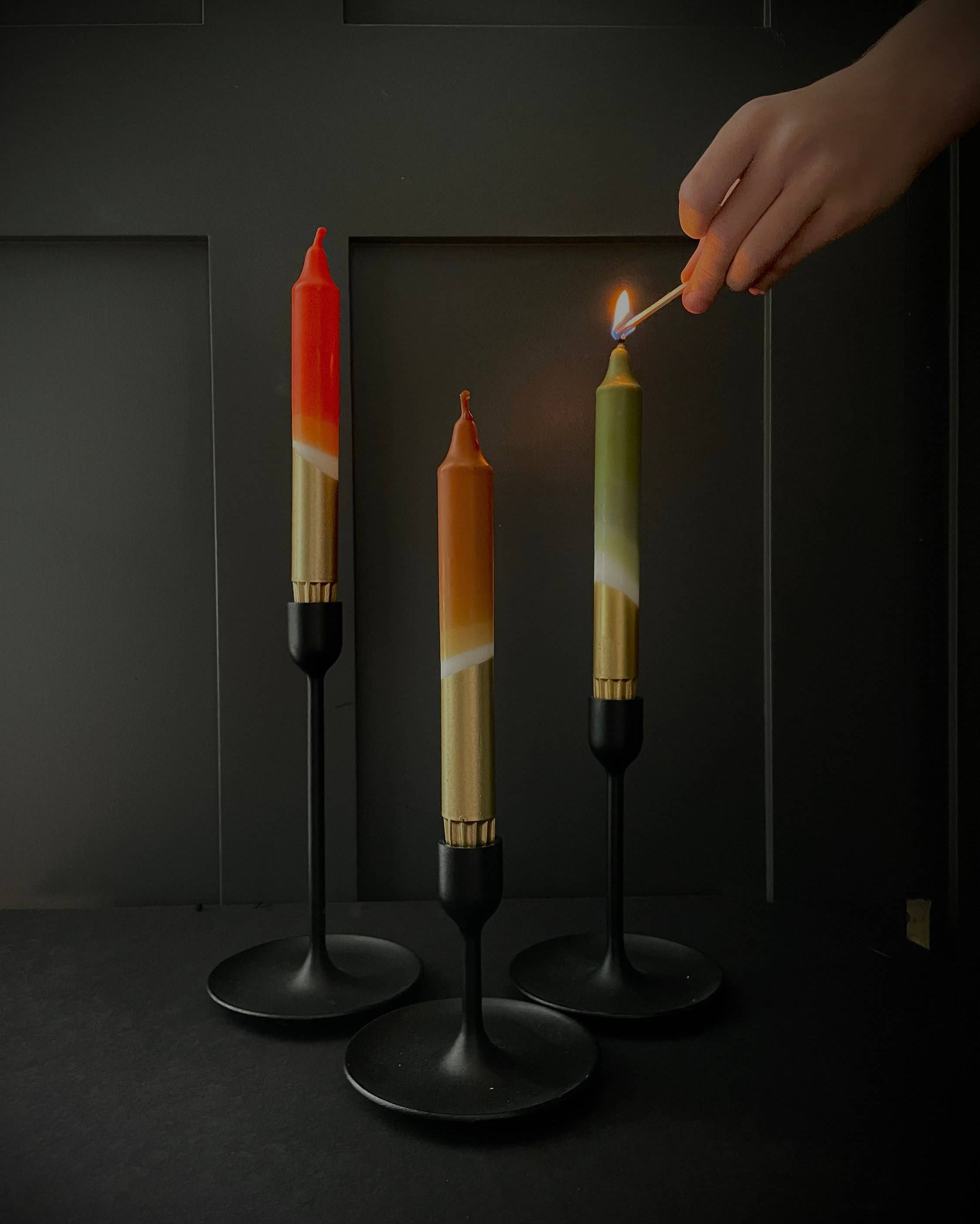 Three colorful candles on black stands with a hand lighting one of them against a dark background.