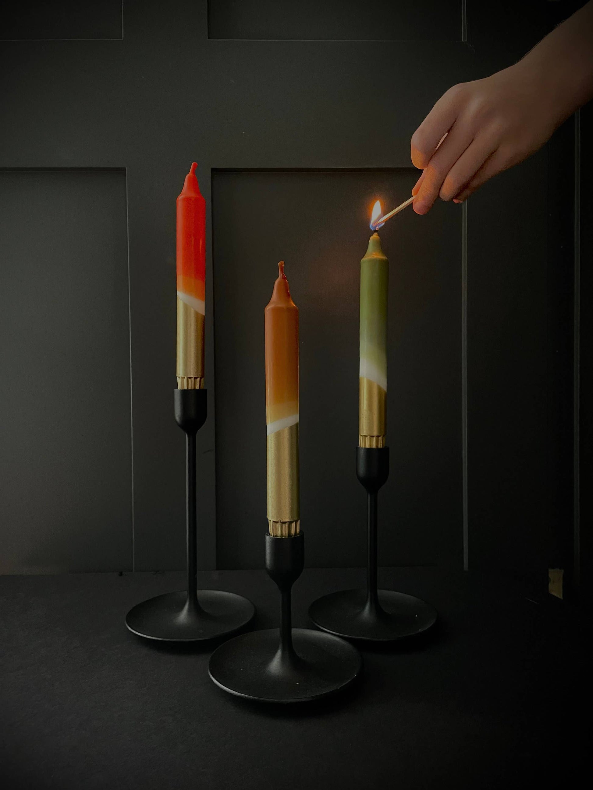 Three colorful candles on black stands with a hand lighting one of them against a dark background.