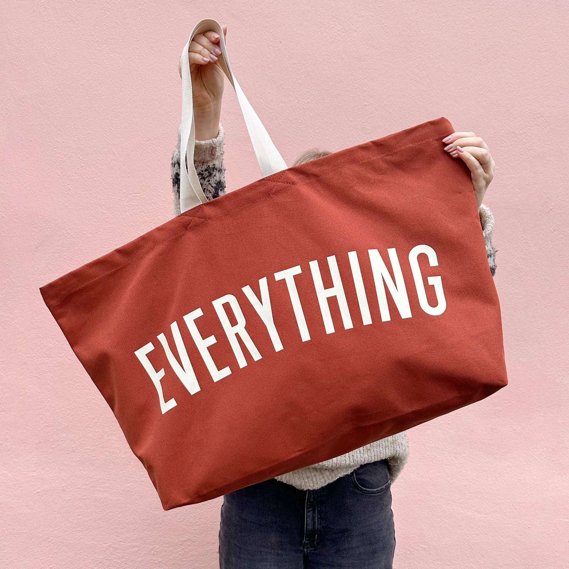 Person holding a red tote bag with 'EVERYTHING' printed on it against a pink background