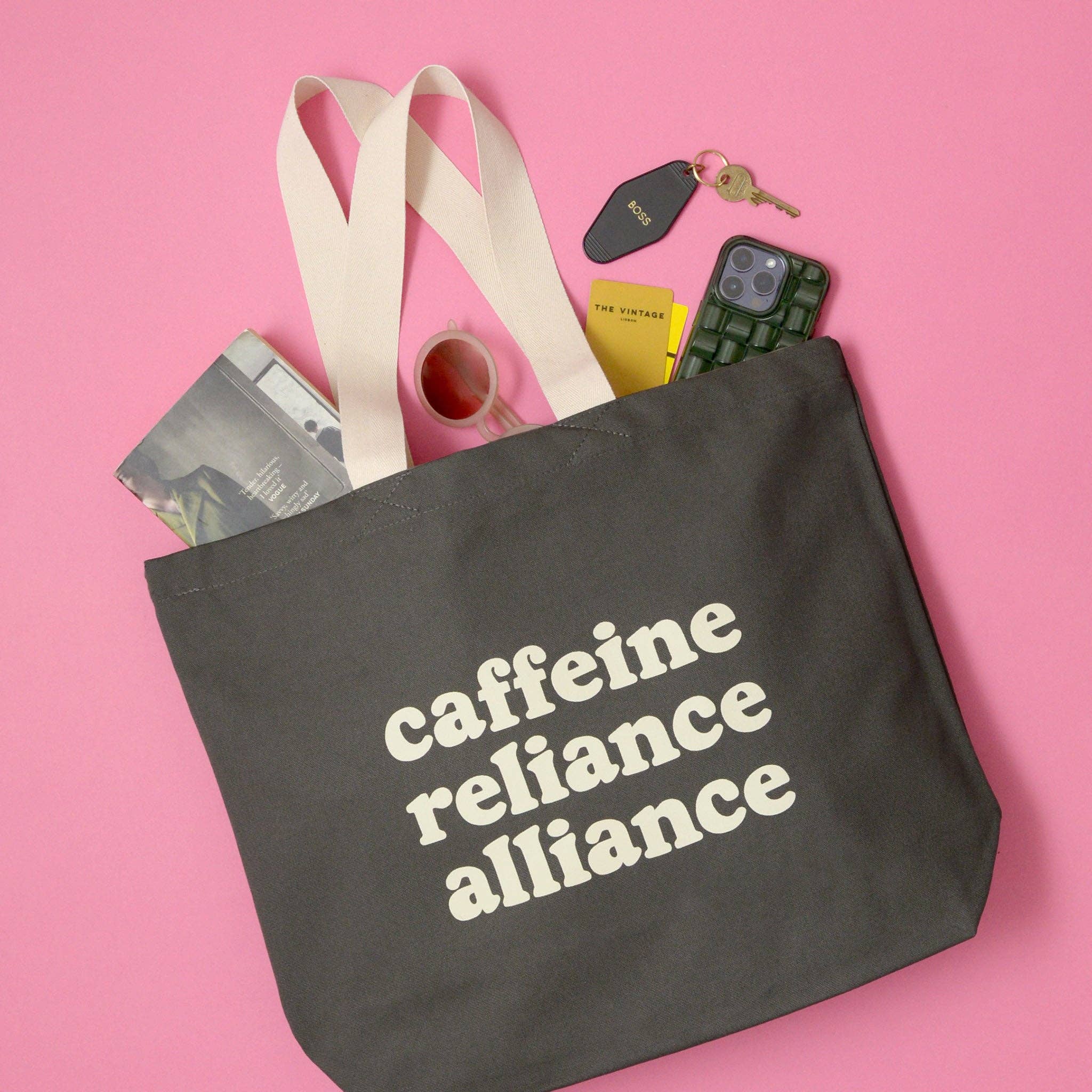 Black tote bag with 'cafeine reliance alliance' text on a pink background