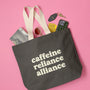 Black tote bag with 'cafeine reliance alliance' text on a pink background