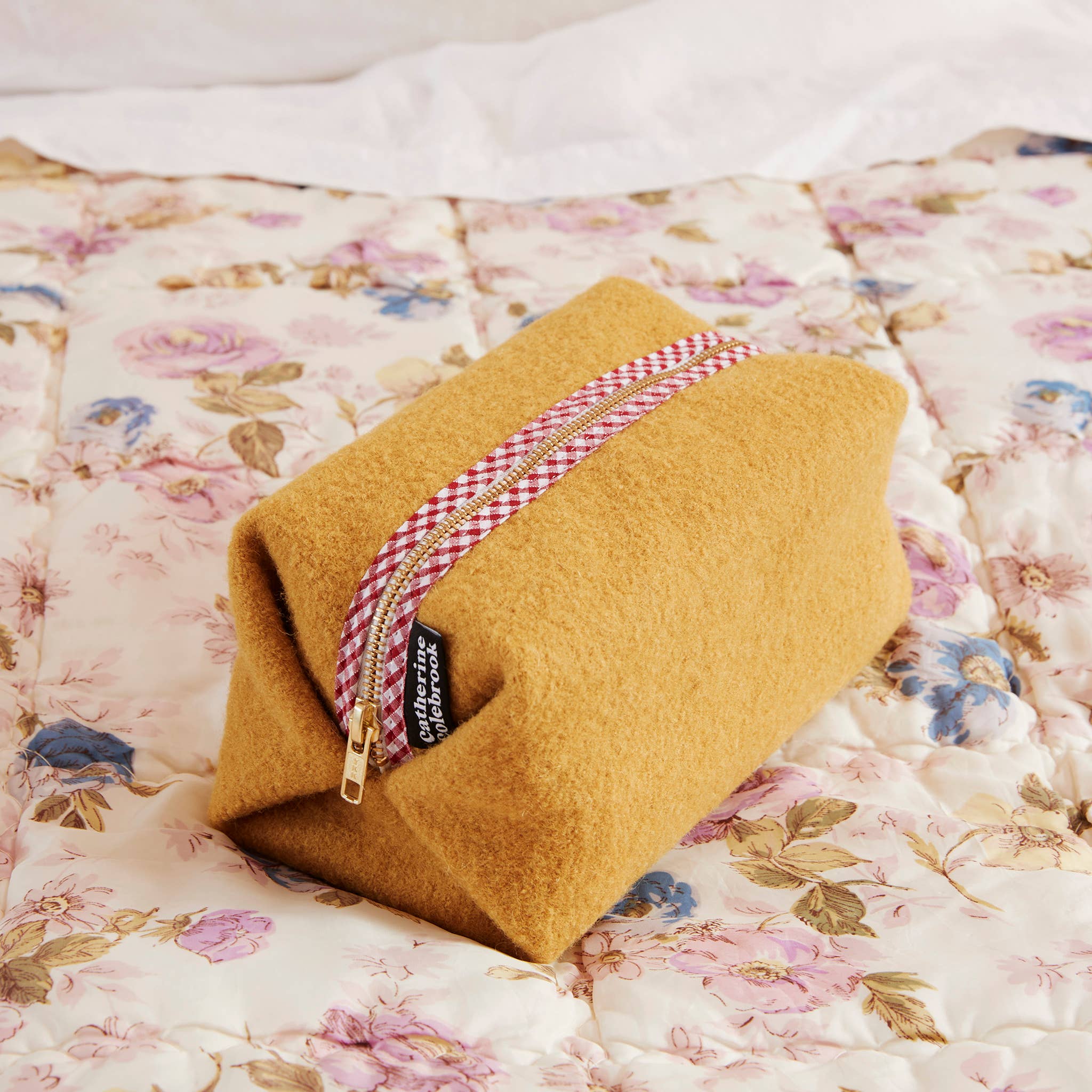Mustard-colored pouch with a red and white checkered zipper on a floral bedspread
