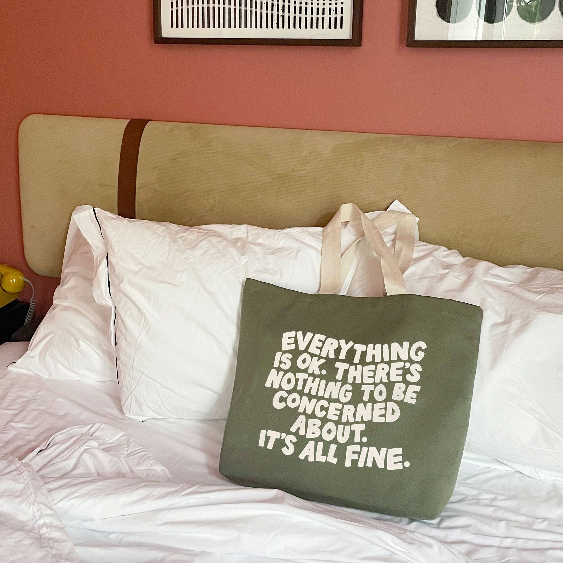 Green tote bag with text on a bed with white pillows and a pink wall.