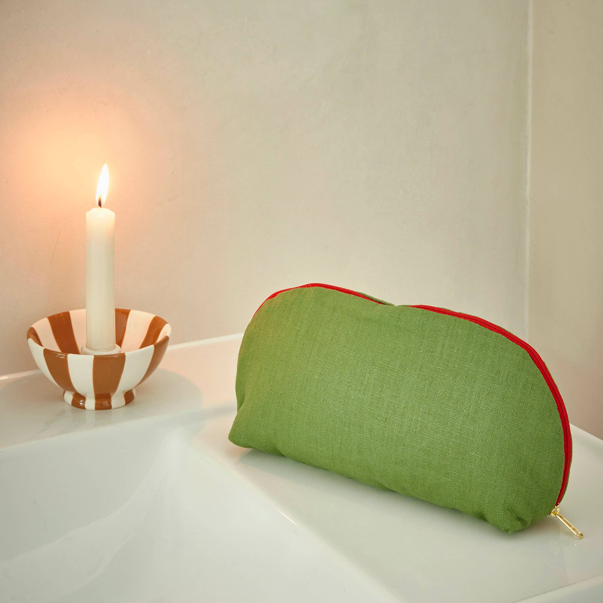 Green cushion with red trim on a white surface next to a striped candle holder with a lit candle.