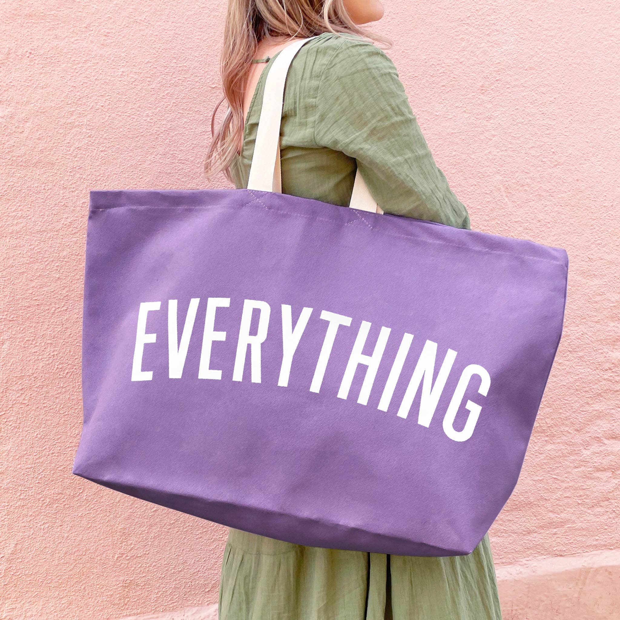 Person holding a purple tote bag with 'EVERYTHING' printed on it against a pink background