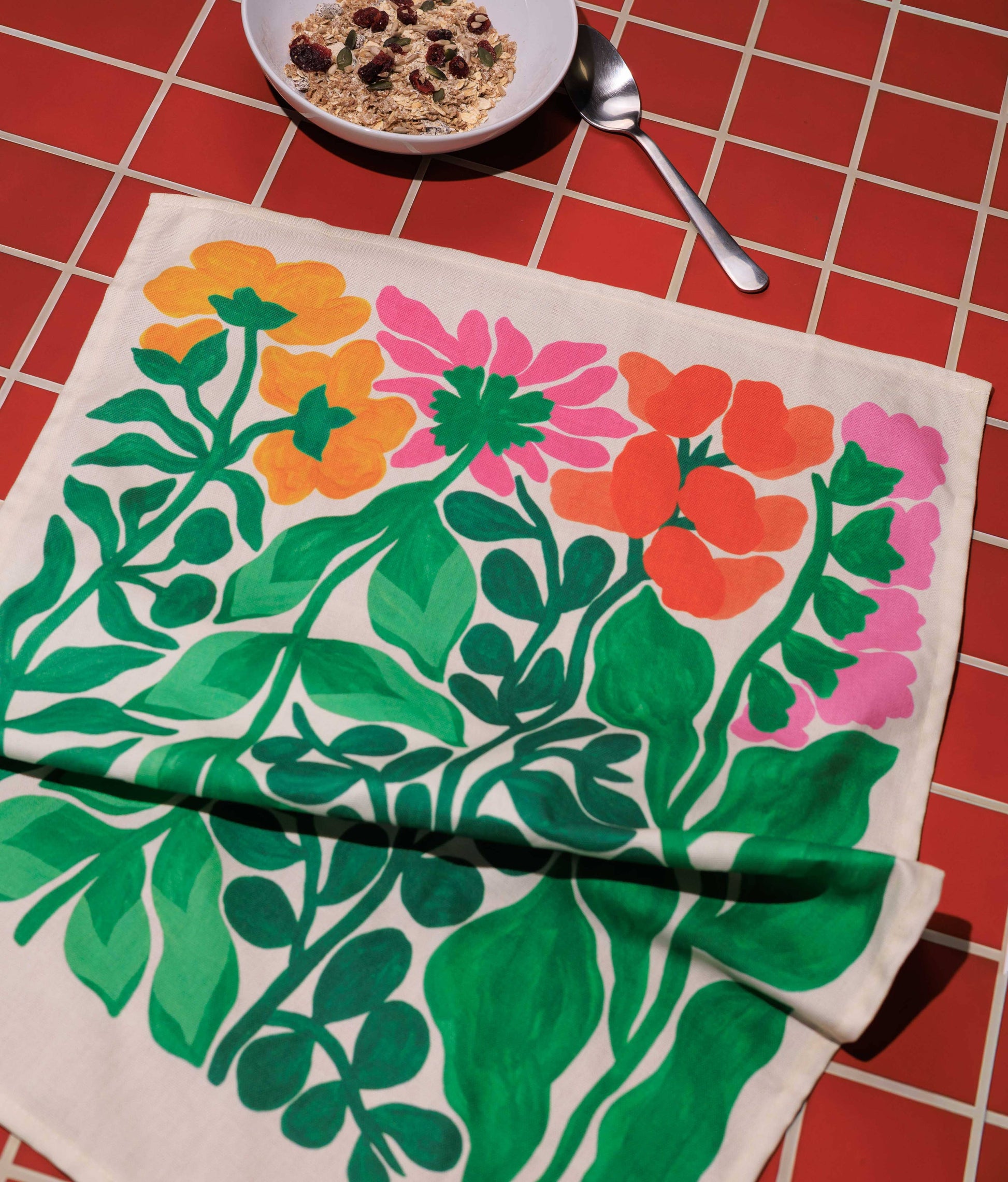 Floral patterned towel on a red checkered tablecloth with a bowl of cereal and spoon.