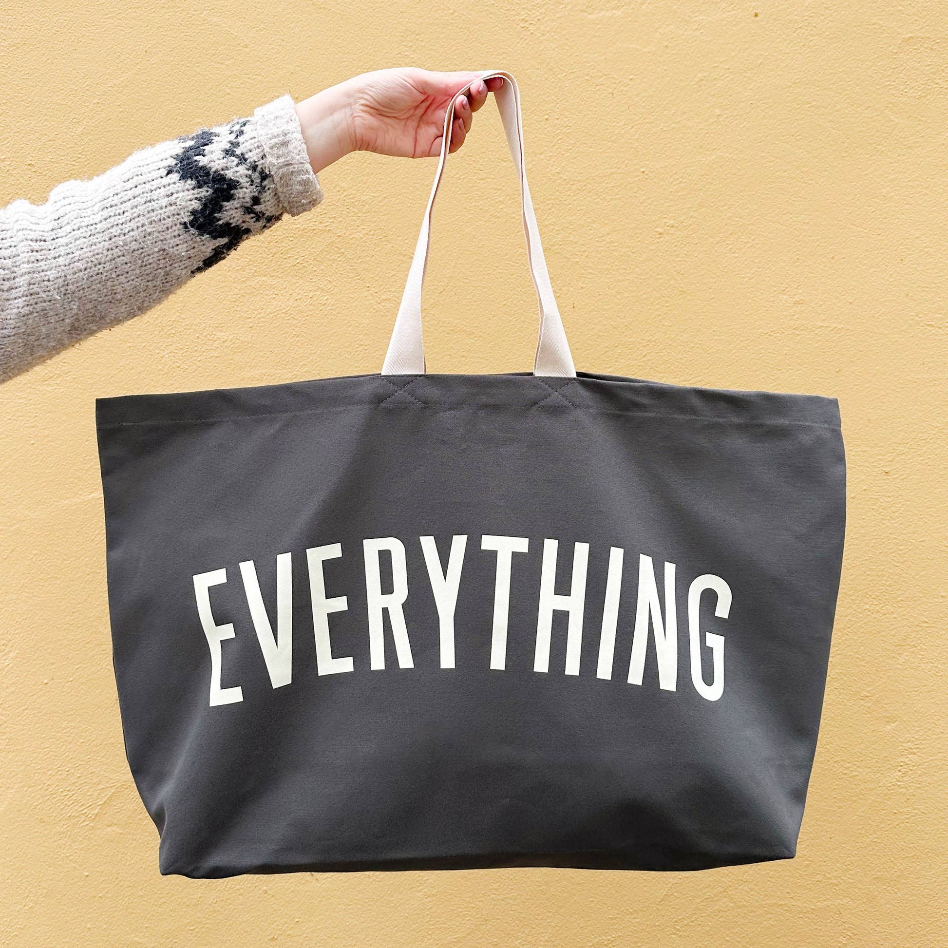 Black tote bag with 'EVERYTHING' text held by a person against a beige background