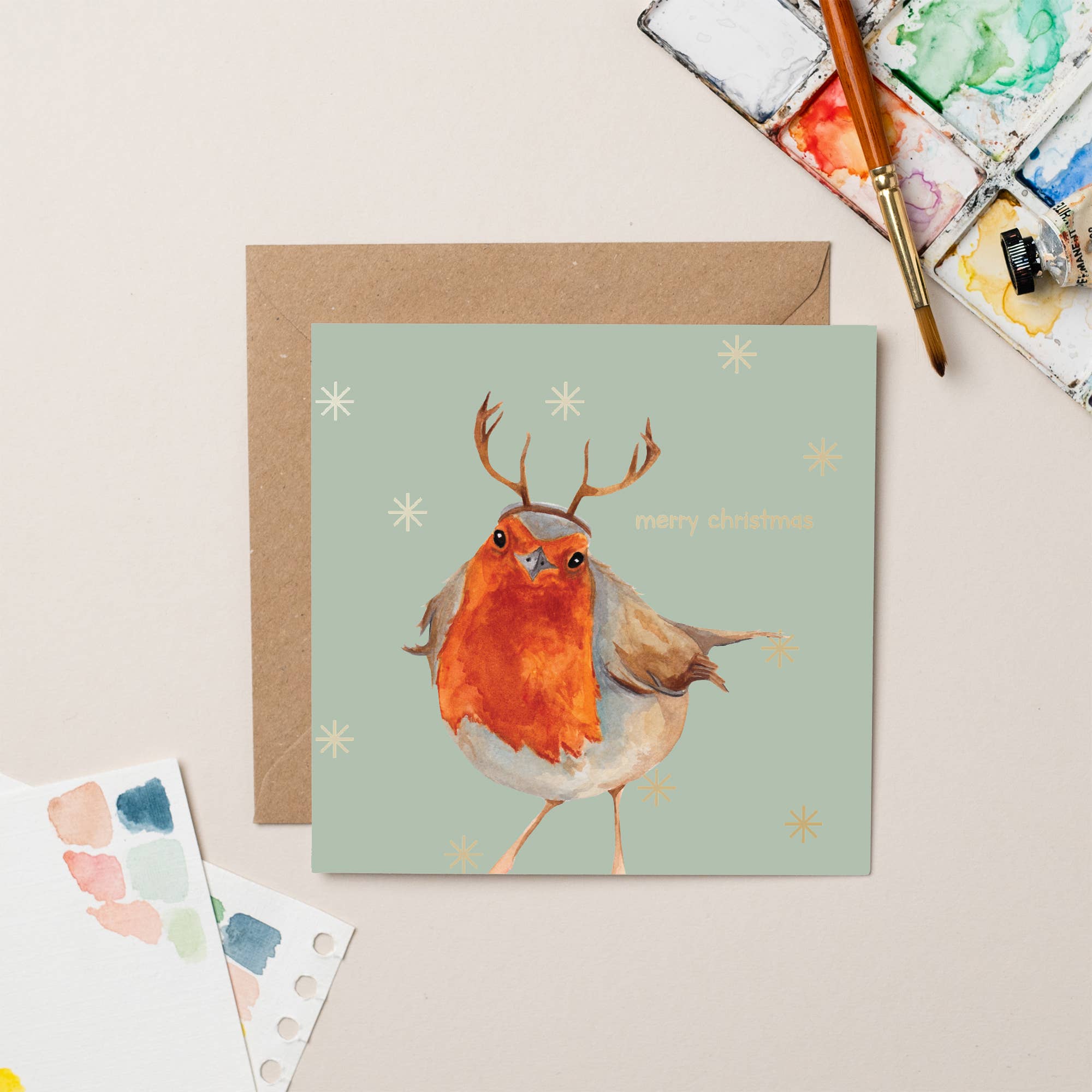 Card with a robin wearing antlers on a light green background, surrounded by watercolor palettes and a brush.