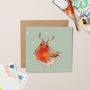 Card with a robin wearing antlers on a light green background, surrounded by watercolor palettes and a brush.