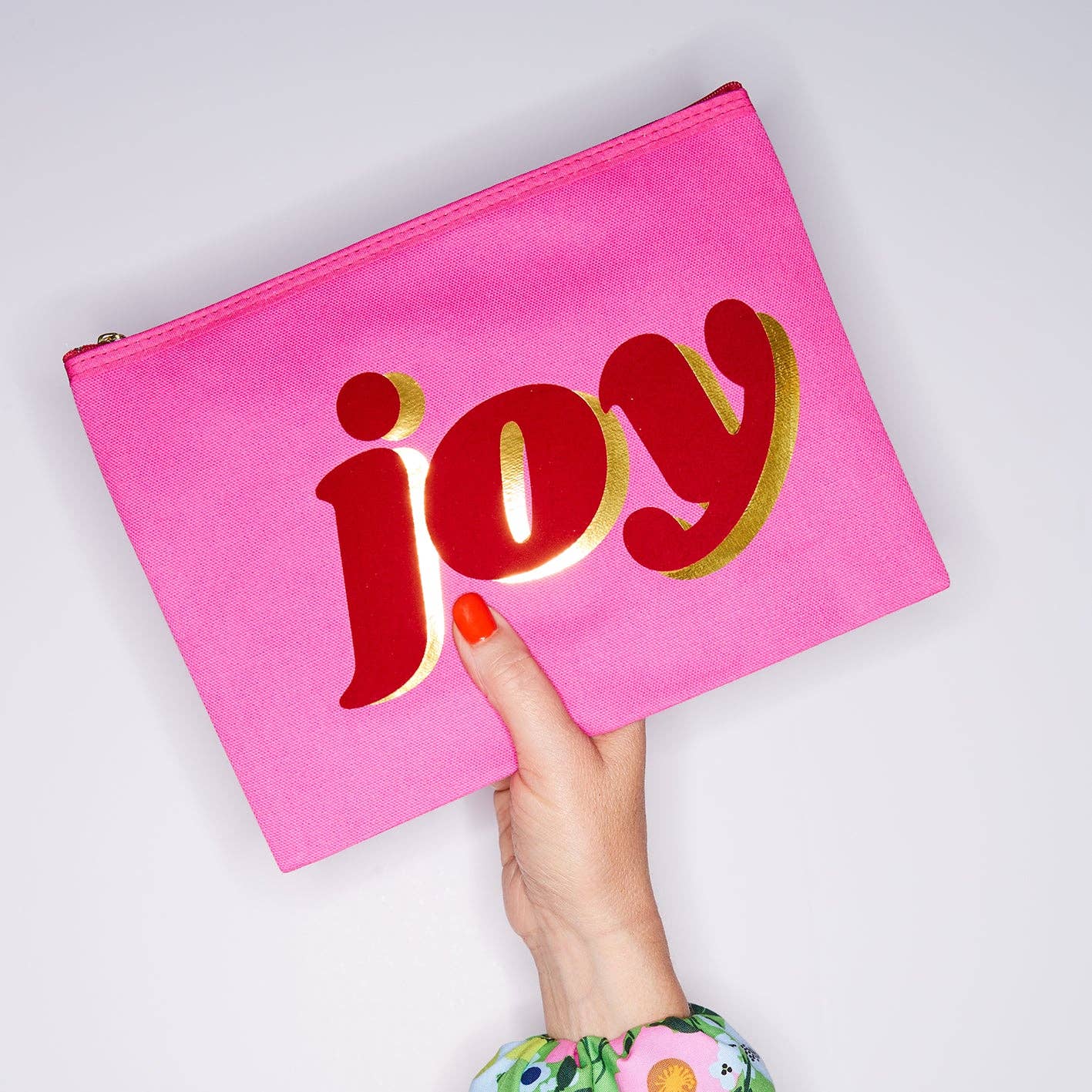 Pink pouch with 'joy' in red and gold held by a hand on a light gray background