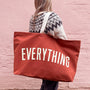 Person holding a red tote bag with 'EVERYTHING' printed on it against a pink brick wall.