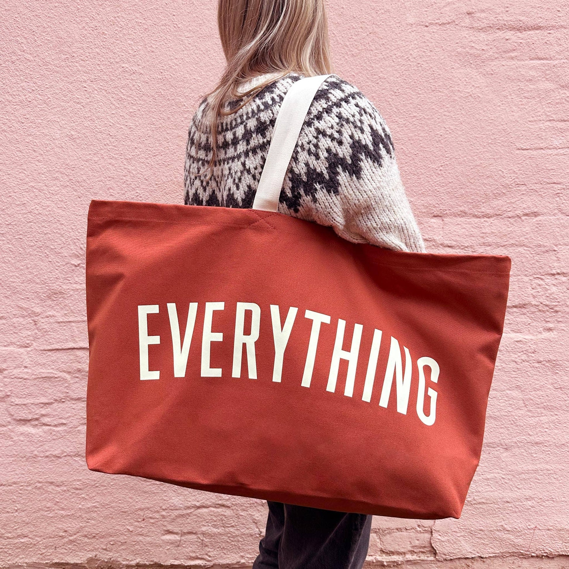 Person holding a red tote bag with 'EVERYTHING' printed on it against a pink brick wall.