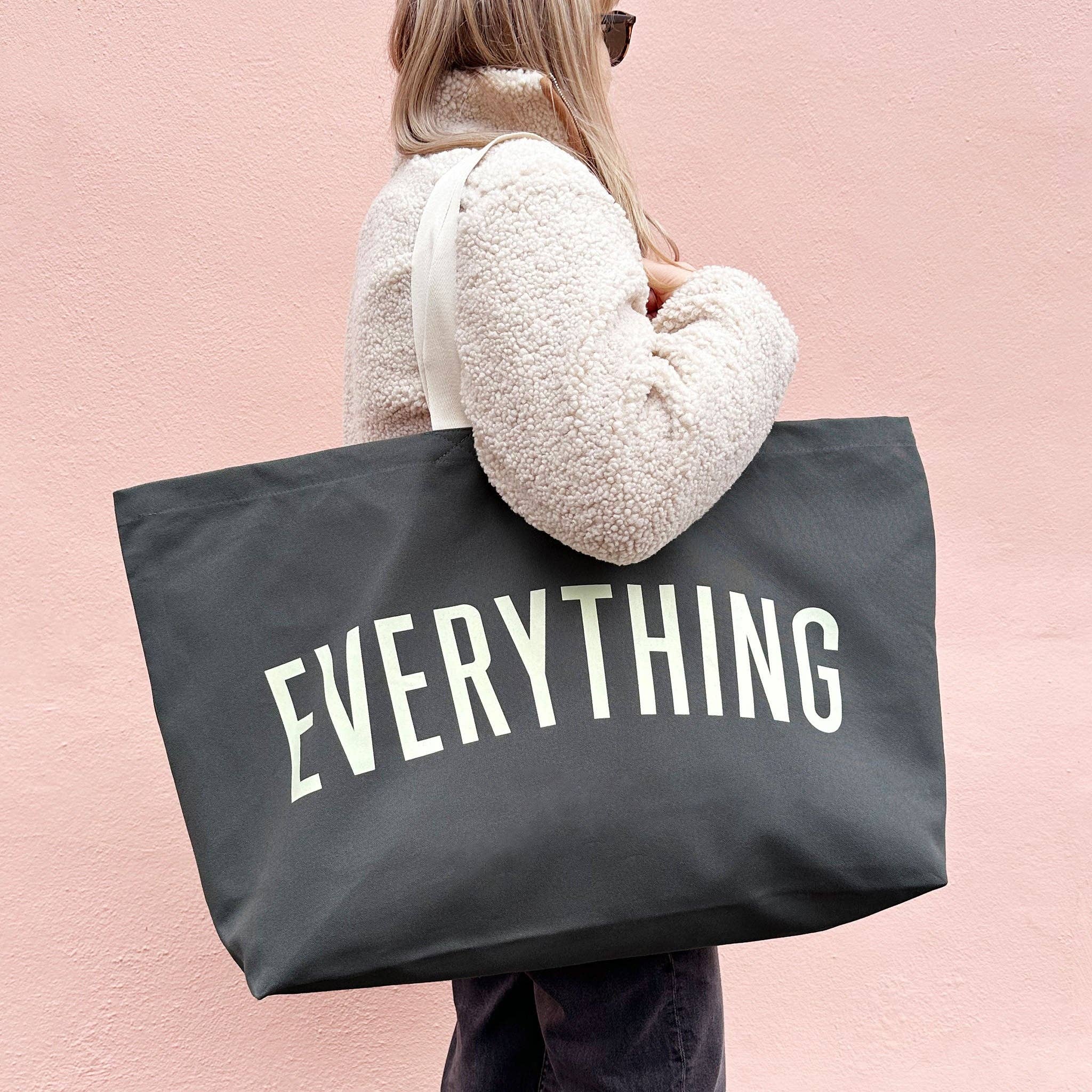 Person holding a black tote bag with 'EVERYTHING' printed on it against a pink background