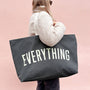 Person holding a black tote bag with 'EVERYTHING' printed on it against a pink background