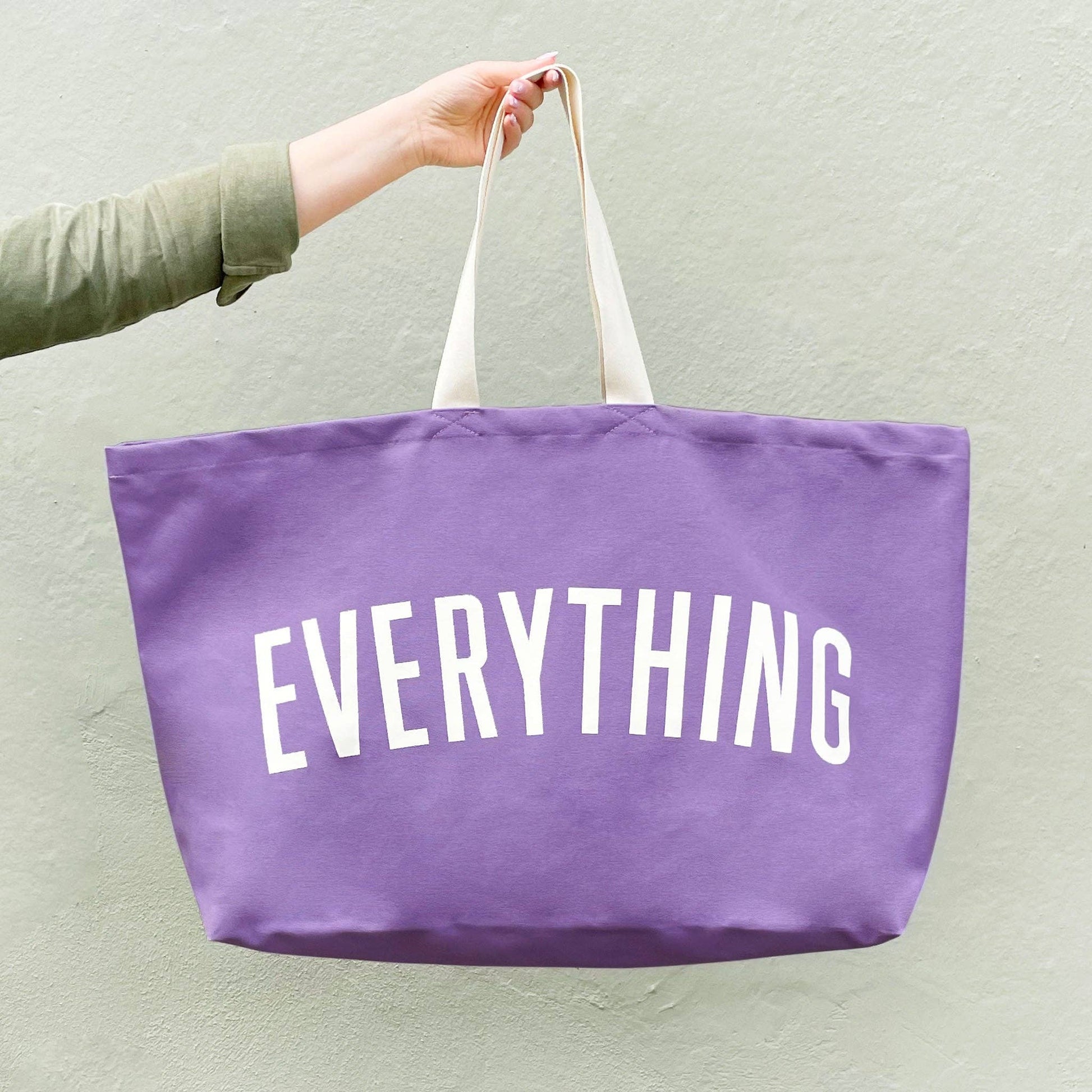 Purple tote bag with 'EVERYTHING' text held by a person against a light green background