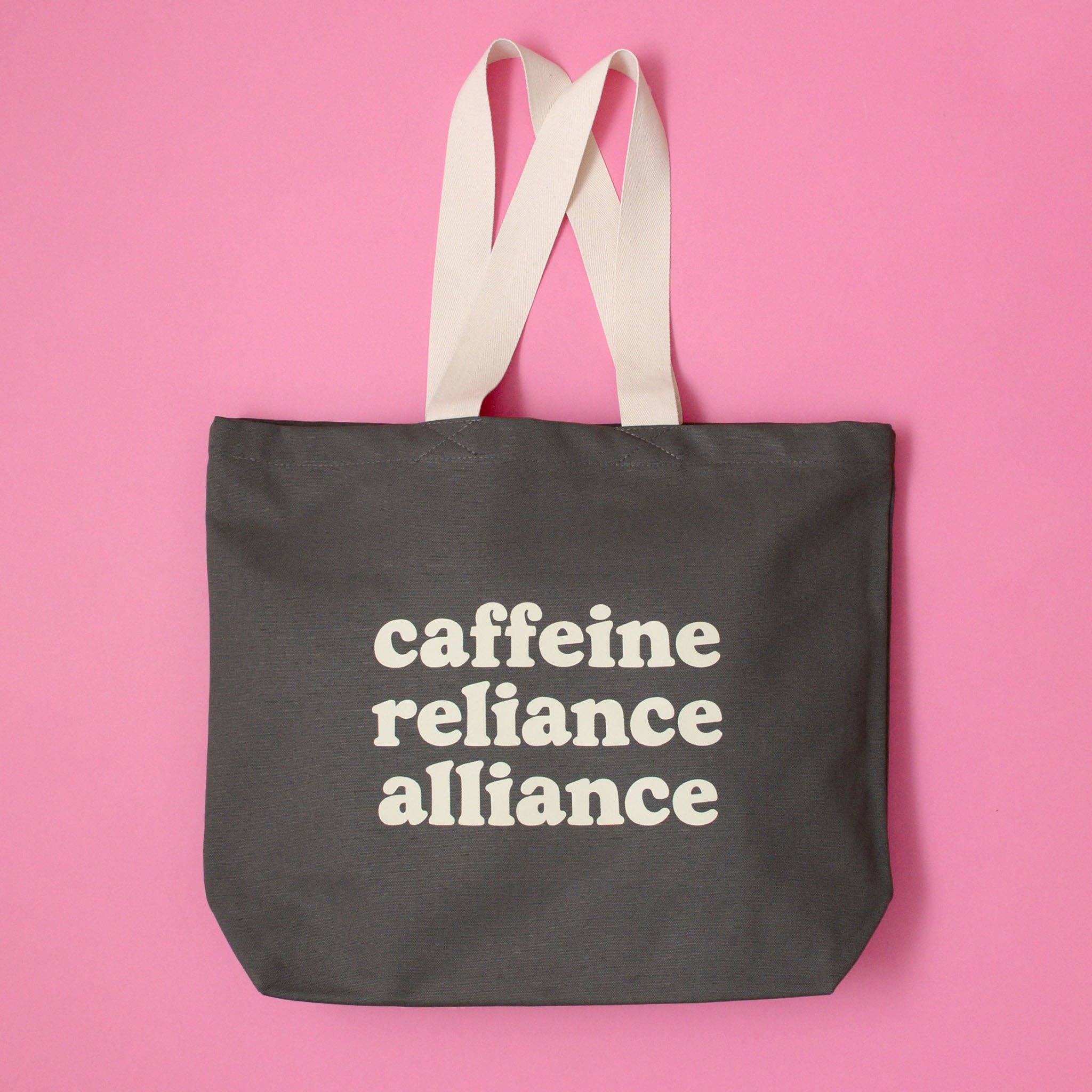 Black tote bag with 'cafeine reliance alliance' text on a pink background