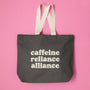Black tote bag with 'cafeine reliance alliance' text on a pink background