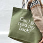 Green tote bag with 'Can I go read my book?' text held by a person wearing a patterned sweater.