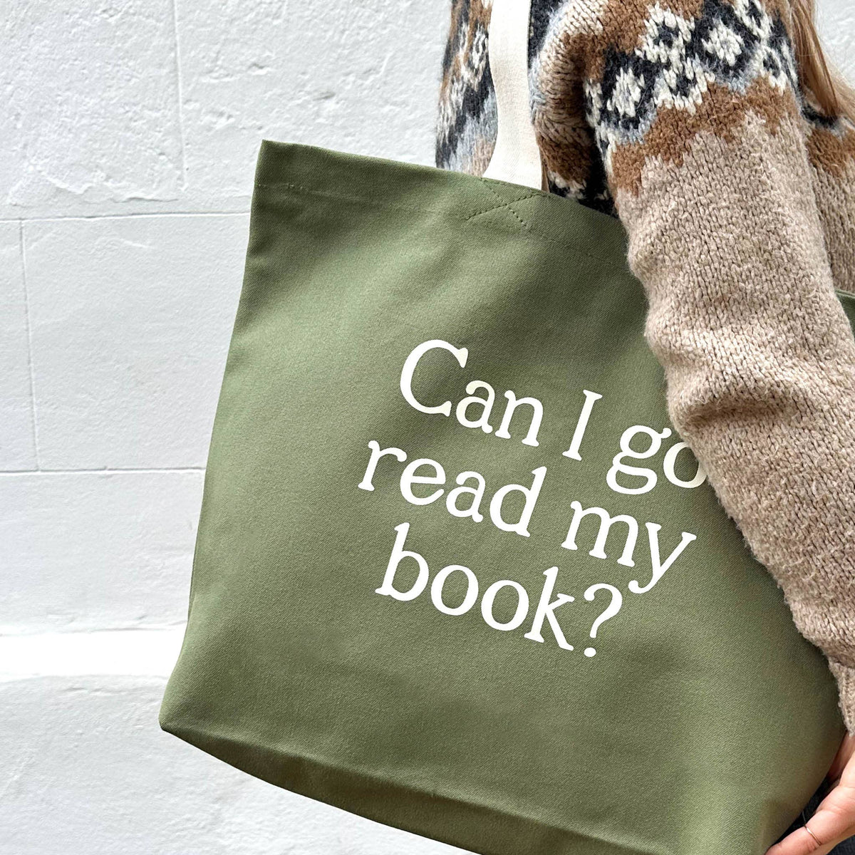 Green tote bag with 'Can I go read my book?' text held by a person wearing a patterned sweater.