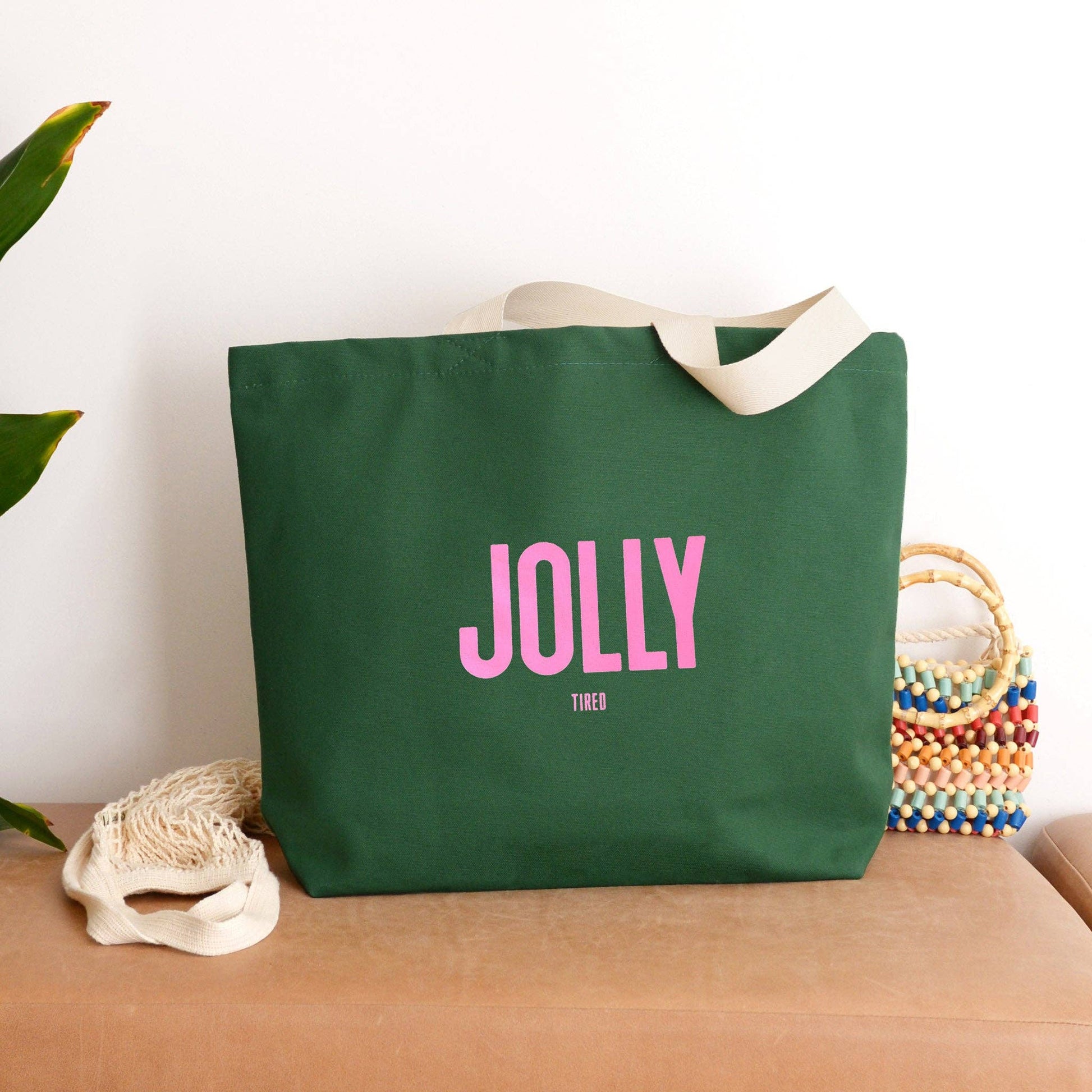 Green tote bag with pink text 'JOLLY' on a wooden surface with a plant and woven basket in the background.