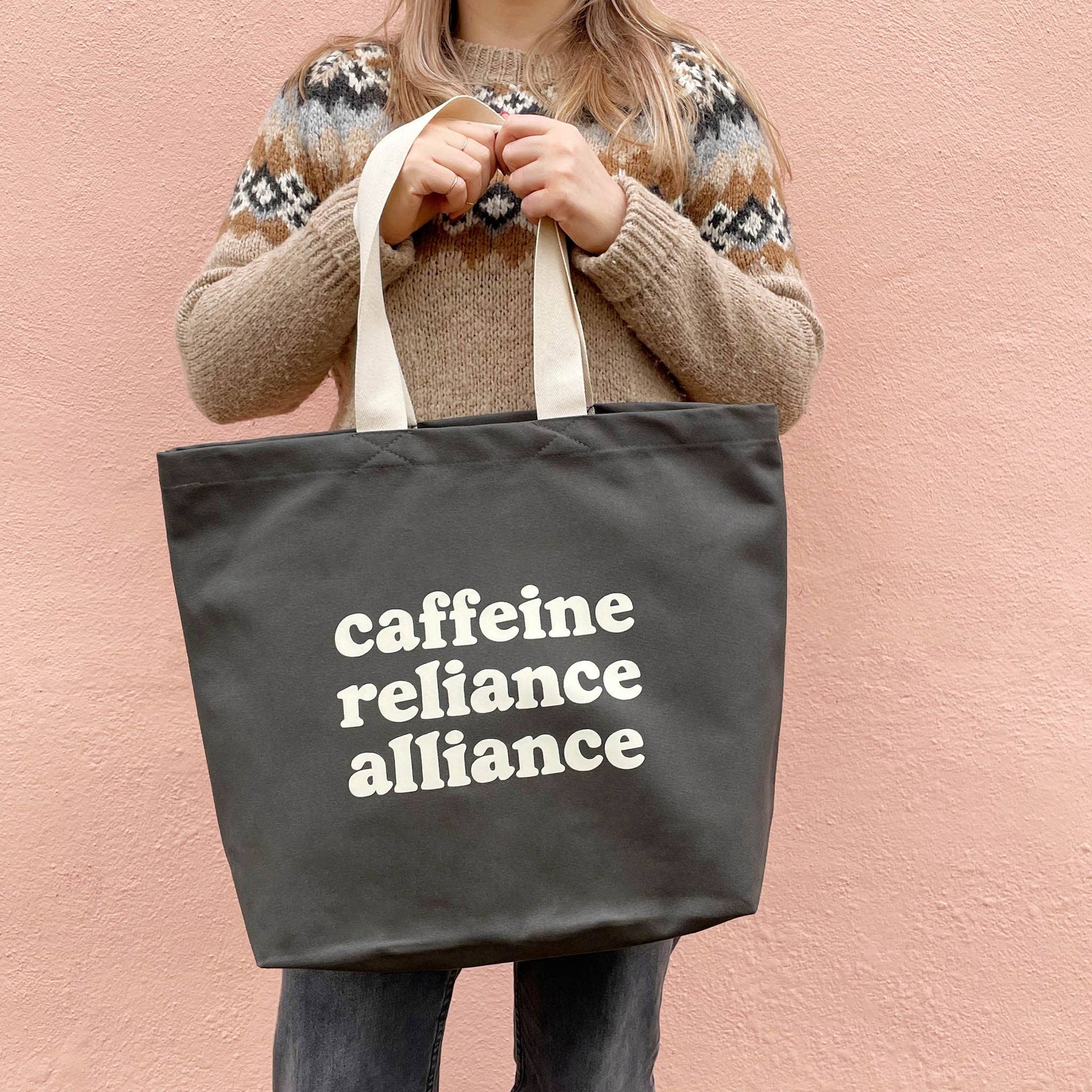 Person holding a tote bag with 'cafeine reliance alliance' text against a pink background
