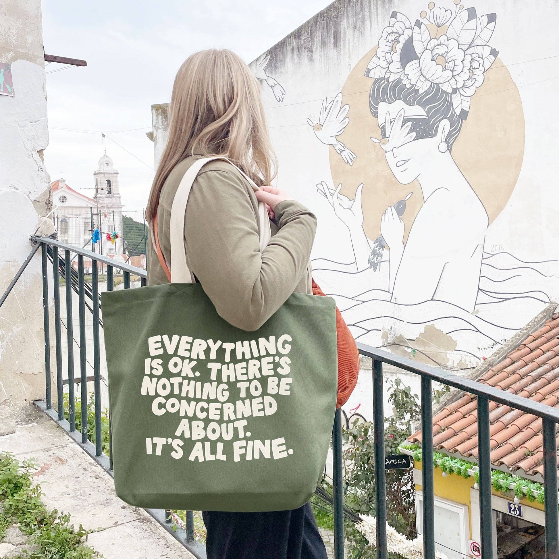Person holding a green tote bag with text, standing on a balcony with a mural in the background.