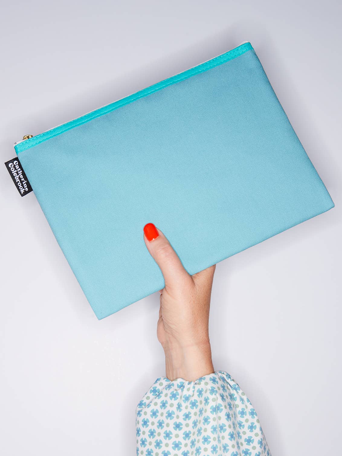 Light blue pouch held by a hand with red nail polish on a light gray background