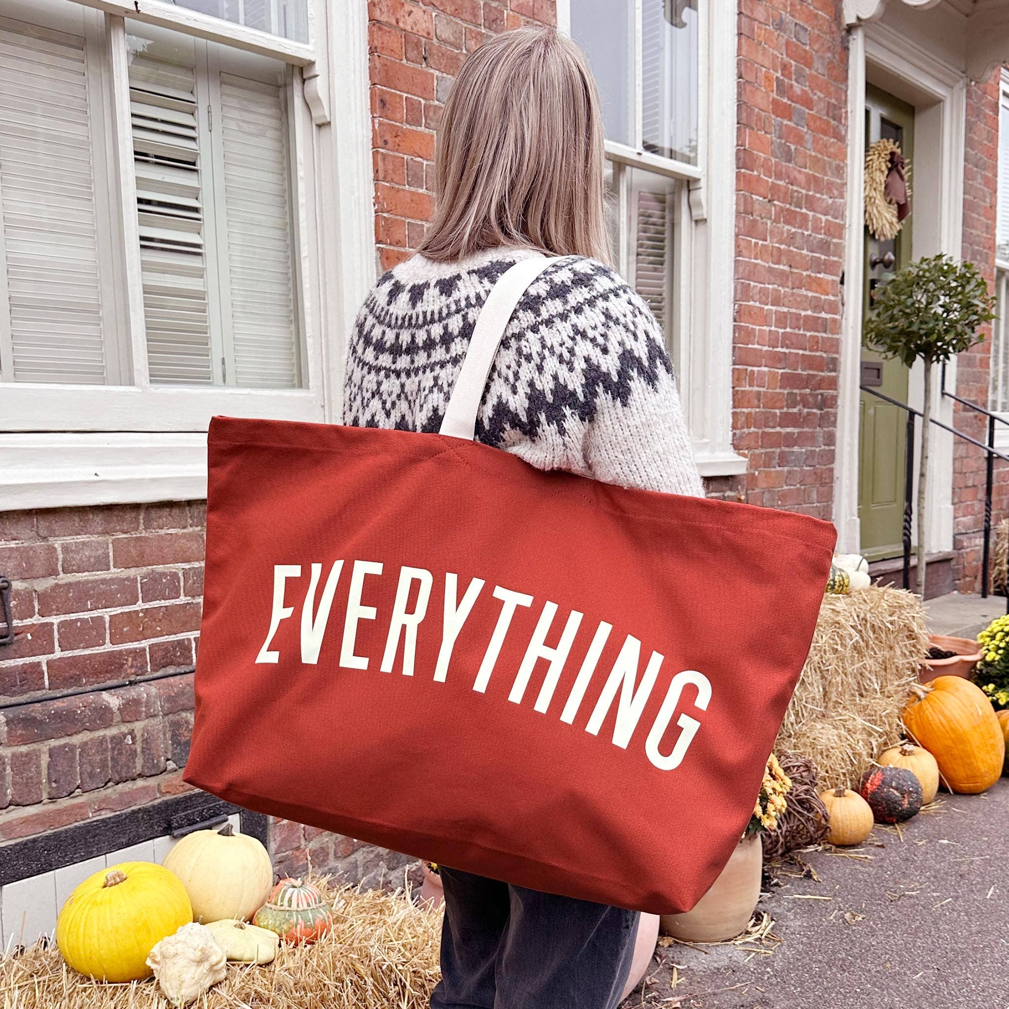 Person holding a red tote bag with 'EVERYTHING' text, standing outside a house with pumpkins and hay.