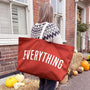 Person holding a red tote bag with 'EVERYTHING' text, standing outside a house with pumpkins and hay.