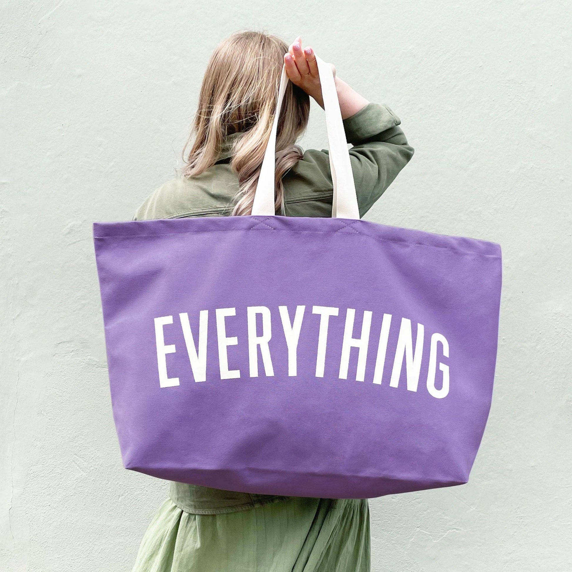 Person holding a purple tote bag with 'EVERYTHING' printed on it against a plain background