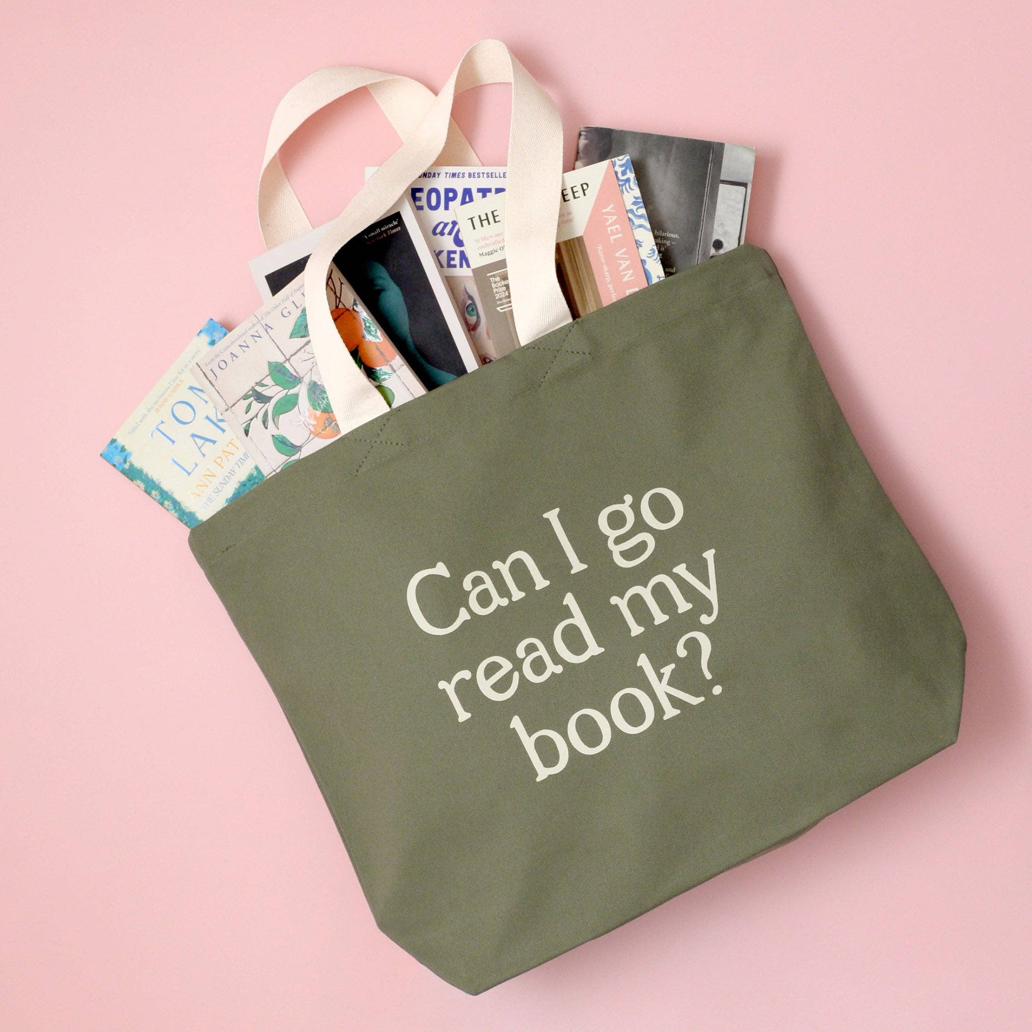 Green tote bag with 'Can I go read my book?' text, filled with books on a pink background