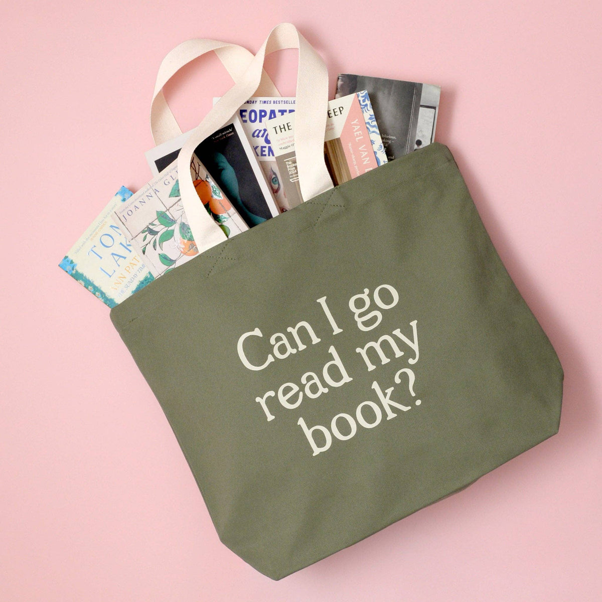 Green tote bag with 'Can I go read my book?' text, filled with books on a pink background