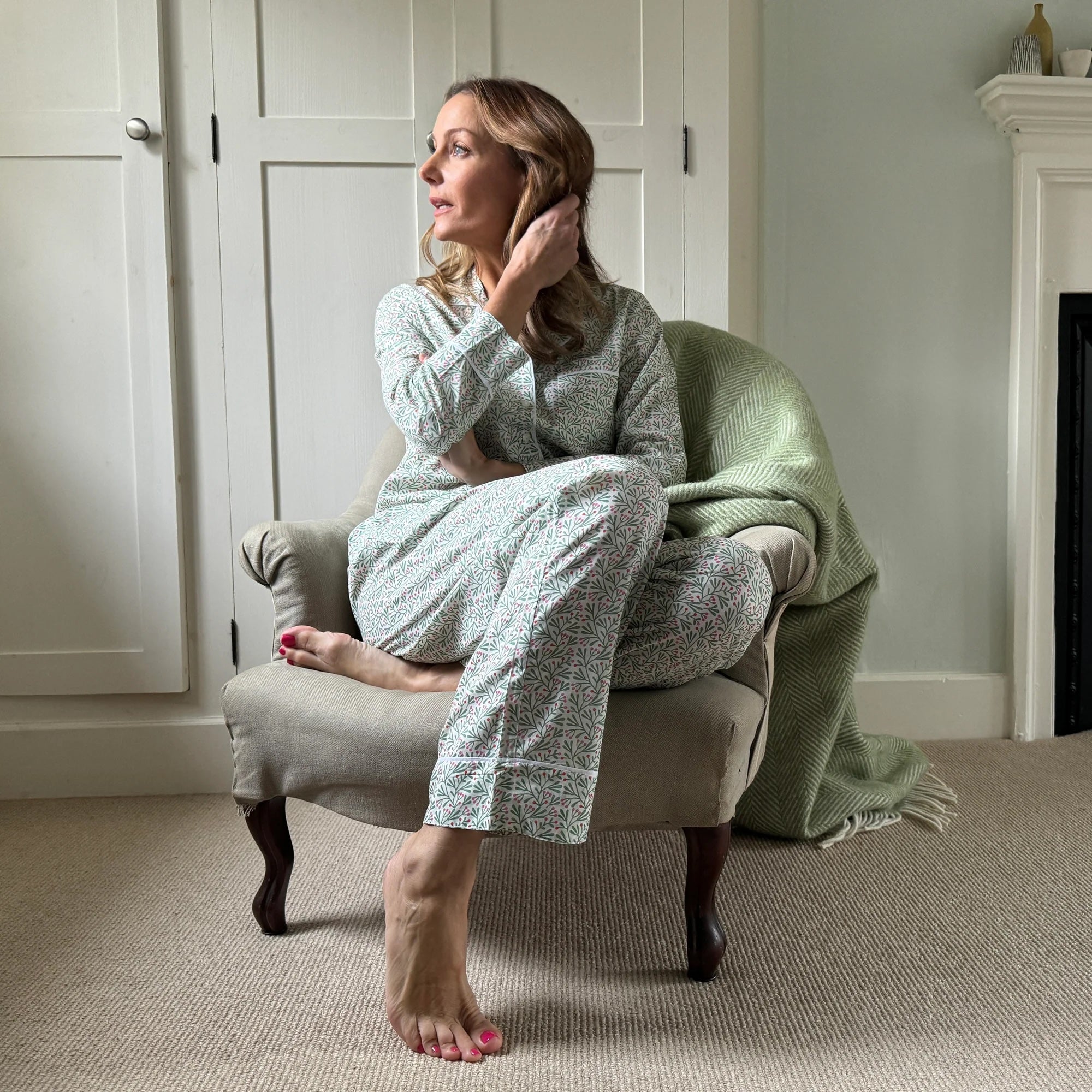 Woman in a cozy room wearing a light green textured robe, sitting on a chair with a green blanket.