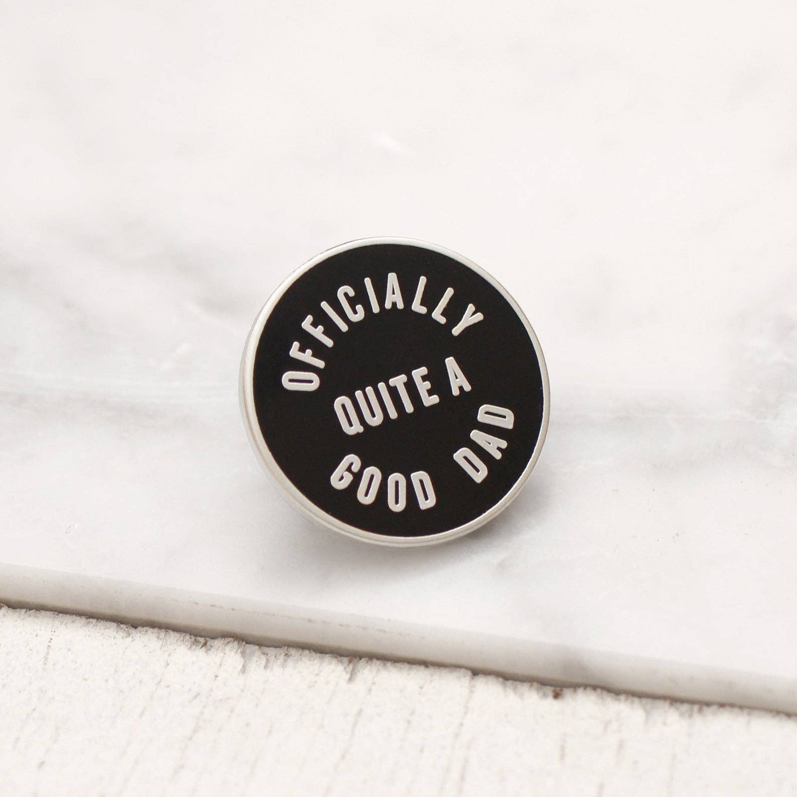 Black and white enamel pin with 'Officially Quite a Good Dad' text on a marble surface.