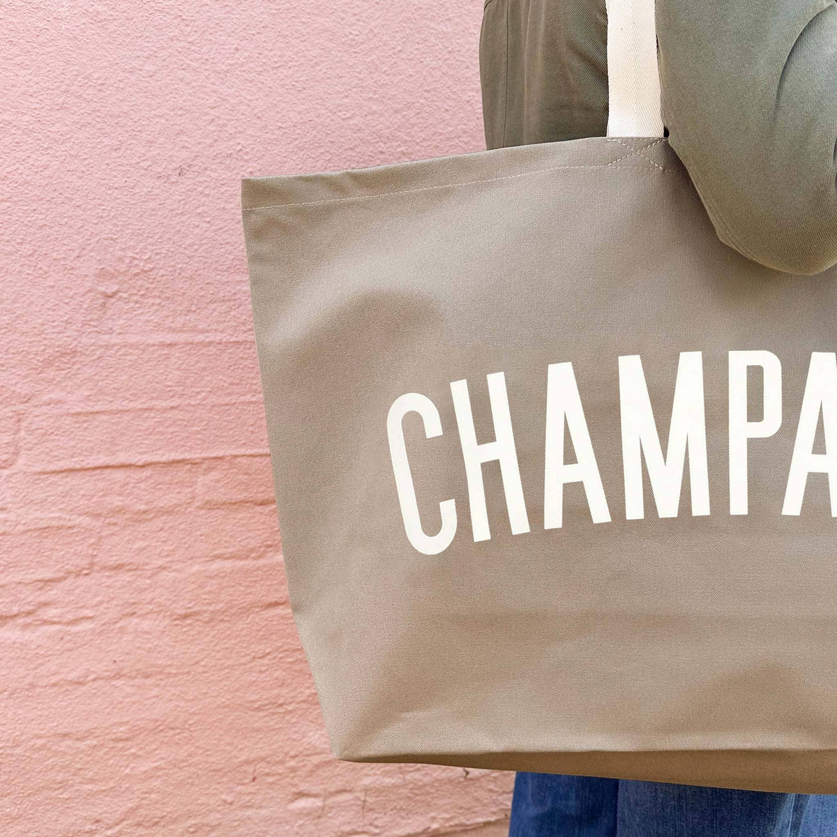 Beige tote bag with 'CHAMPA' text held by a person against a pink wall.