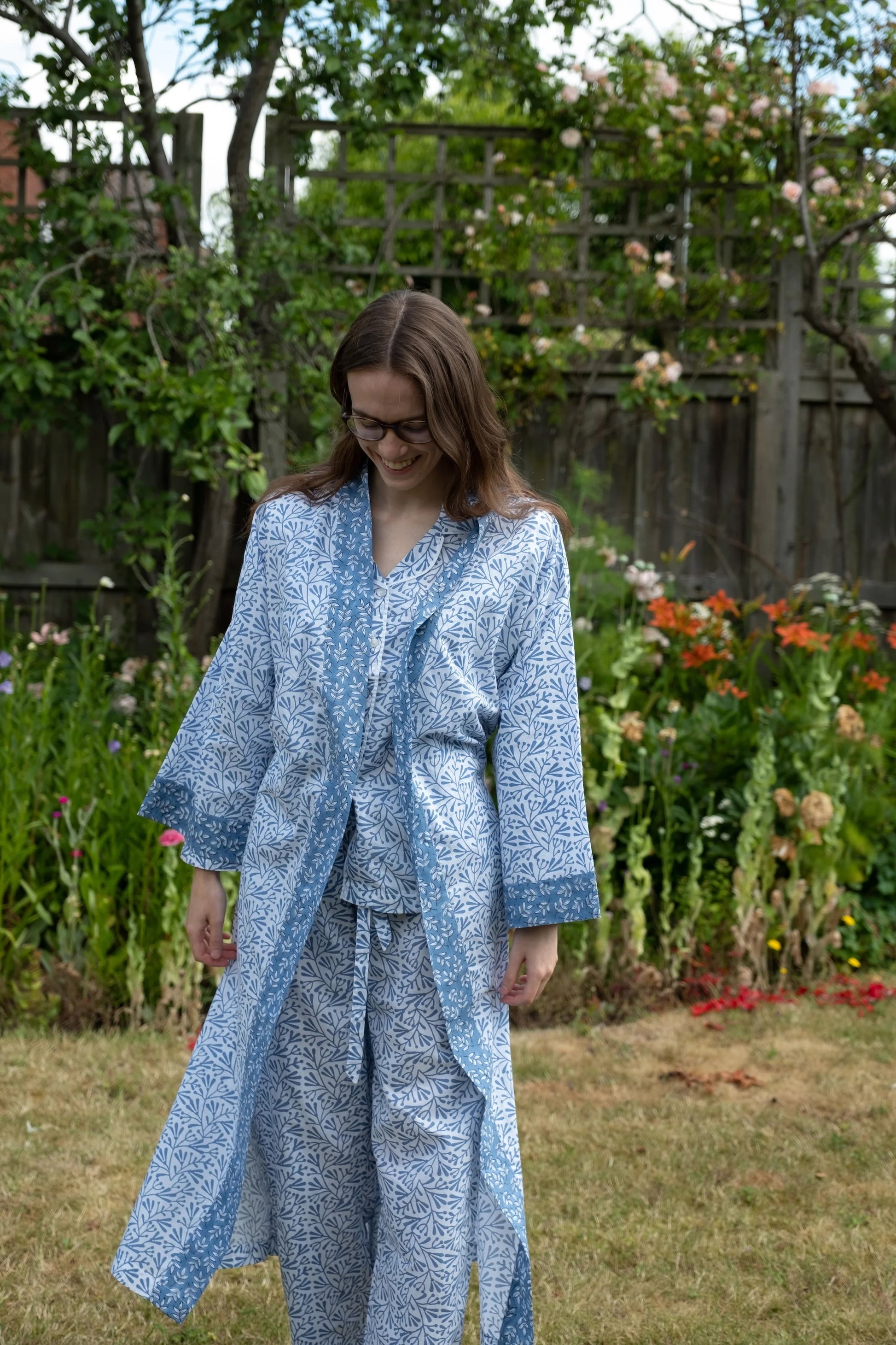 Woman in a blue patterned outfit standing in a garden with flowers and greenery.