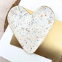 Heart-shaped ceramic plate with multicolored speckles on a gold base, placed on a white surface.