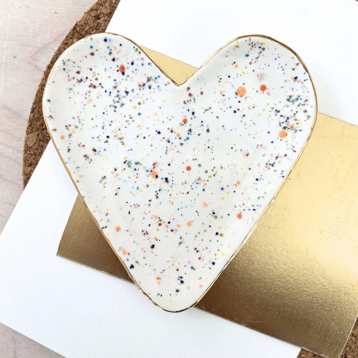 Heart-shaped ceramic plate with multicolored speckles on a gold base, placed on a white surface.