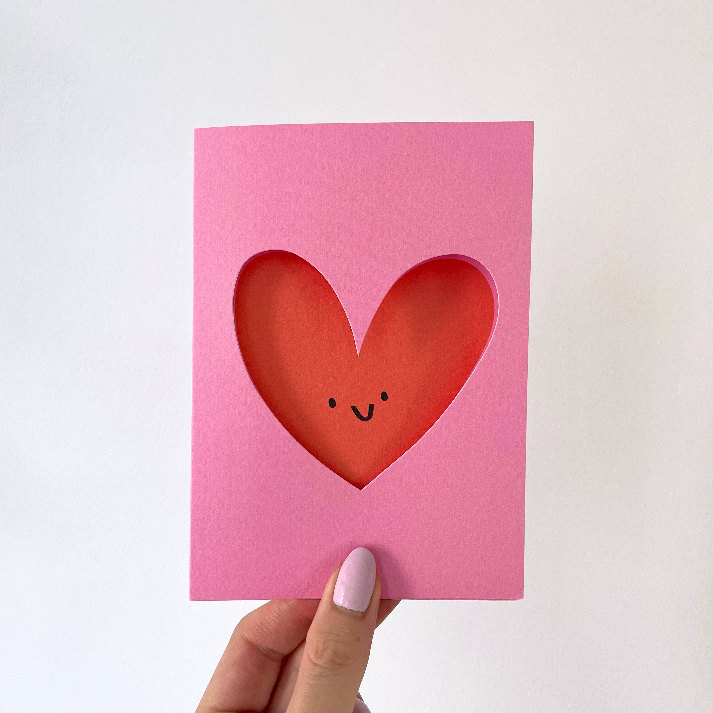 Pink card with a red heart cutout featuring a smiley face, held by a hand on a white background.