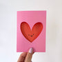 Pink card with a red heart cutout featuring a smiley face, held by a hand on a white background.
